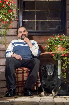 На картине изображены мужчина и черная собака. Мужчина сидит на стуле на клетчатом пледе. Зеленая листва с красными цветочками, стиль Под масло, художник Виктория Б
