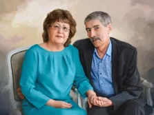 Парный портрет под масло. На картине изображён парный портрет: женщина в бирюзовом платье и очках сидит рядом с мужчиной в тёмном пиджаке и голубой рубашке. Они держатся за руки, их позы спокойны и доверительны, а фон выполнен в мягких, размытых тонах, создающих тёплую атмосферу. Художник Анастасия