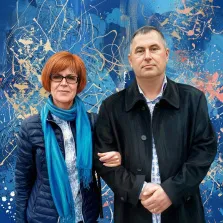 Парный портрет в стиле дрим арт. На портрете изображены мужчина и женщина: женщина с короткими рыжими волосами, в очках, тёмно-синей куртке и ярком голубом шарфе слегка улыбается, а мужчина в чёрном пальто и рубашке с узором стоит рядом, сложив руки перед собой с серьёзным выражением лица. Фон выполнен в абстрактной манере с преобладанием синих оттенков и хаотичных мазков белого, жёлтого и красного цветов, создающих динамичную атмосферу композиции. Художник: Ирина