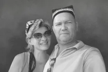 Парный портрет в стиле карандаш. На портрете изображены мужчина и женщина: мужчина в светлой рубашке и традиционной шапке с орнаментом, с серьёзным выражением лица; женщина в солнечных очках и платке с узором, с тёплой улыбкой, слегка наклонившаяся к мужчине. Фон нейтральный, однотонный, подчёркивает фигуры героев и создаёт ощущение близости между ними. Художник: Антонина