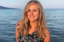 Женский портрет в стиле под масло. На портрете изображена молодая женщина с длинными светлыми волнистыми волосами, одетая в чёрное платье с цветочным принтом; она тепло улыбается, её взгляд открыт и доброжелателен. На фоне — спокойное море с мягкими волнами и приглушённый горизонт, создающий атмосферу умиротворения и лёгкости. Художник: Александра 