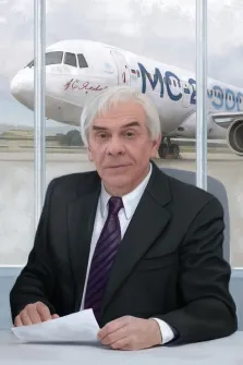 Мужской портрет под масло. На портрете изображён пожилой мужчина с седыми волосами, в тёмном костюме, белой рубашке и фиолетовом галстуке; он сидит за столом, держа в руках лист бумаги, с серьёзным и сосредоточенным выражением лица. На заднем плане виден самолёт МС-21 с надписью «А.С. Яковлев» на фюзеляже, что придаёт композиции деловой и индустриальный характер. Художник: Антонина