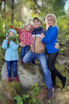 Семейный портрет под масло. На портрете изображена счастливая семья из четырёх человек на фоне зелёного леса: отец в сине-коричневой куртке и кепке сидит на камне, обнимая одной рукой дочь в яркой розовой куртке, рядом стоит мать в синей куртке и чёрных брюках, а младшая дочь в голубой куртке держит в руках ветку с красными ягодами. Солнечные лучи, пробивающиеся сквозь листву, создают тёплую и радостную атмосферу семейного единения на природе. Художник: Антонина