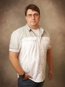 Мужской портрет под масло. На портрете изображён мужчина в очках с короткими каштановыми волосами; он одет в светлую рубашку с короткими рукавами поверх белой футболки, тёмные джинсы, на левом запястье — чёрные часы. Мужчина стоит в расслабленной позе, одна рука в кармане, выражение лица спокойное, с лёгкой улыбкой, фон — нейтральный, текстурированный, серо-бежевого оттенка. Художник: Антонина