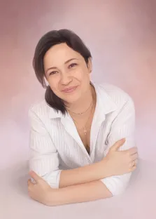 Женский портрет под масло. На портрете изображена женщина с короткими тёмными волосами, в белой рубашке в тонкую полоску; она слегка наклонилась вперёд, скрестив руки, и тепло улыбается, глядя в камеру. Нежный розово-бежевый фон подчёркивает мягкость и доброжелательность её образа, а изящные украшения добавляют утончённости. Художник: Антонина