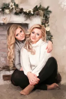 Семейный портрет под масло. На портрете изображены две женщины с длинными светлыми волосами — вероятно, мать и дочь: одна в белом свитере и чёрных брюках сидит, скрестив ноги и мягко улыбаясь, другая, в сером свитере, обнимает её за плечи, слегка наклонившись вперёд с тёплой улыбкой. На фоне — украшенный еловыми ветками и шишками камин, создающий уютную, праздничную атмосферу. Художник: Александра 