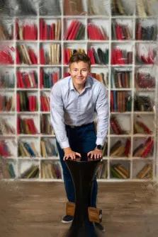 Мужской портрет под масло. На портрете изображён молодой человек в светло-голубой рубашке и тёмно-синих брюках, который с лёгкой улыбкой опирается руками на чёрный стол необычной формы. На фоне — книжный стеллаж, заполненный книгами с красными и тёмными корешками, создающий атмосферу библиотеки или кабинета. Художник: Александра 