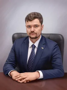 Под масло, художник Антонина На изображении представлен портрет мужчины, сидящего за столом. У мужчины короткая стрижка каштанового цвета и небольшая борода. Он одет в темно-синий костюм, белую рубашку и галстук с узором. На лацкане пиджака закреплен небольшой значок золотистого цвета. Под масло, художник Антонина