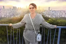 На изображении женщина в платье с геометрическим рисунком, стоит на балконе с синими перилами. Она держится за перила руками. На ней надеты очки в прямоугольной оправе, а также украшения: ожерелье с кулоном и сумка через плечо. Волосы убраны в прическу. Под масло, художник Артём