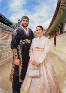 Парный портрет под масло. На картине изображены мужчина и женщина в традиционных корейских нарядах — ханбоках: мужчина одет в тёмный ханбок с золотыми узорами и головным убором, женщина — в нежно-розовый ханбок с золотой вышивкой, держа в руках небольшую сумочку. Они стоят на фоне традиционной корейской архитектуры с черепичными крышами и кирпичными стенами, на фоне ясного неба с облаками. Художник Анна