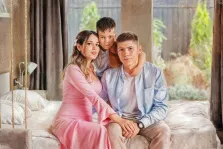 Семейный портрет в стиле под масло. Женщина в нежно-розовом платье сидит рядом с молодым мужчиной в голубой рубашке, а позади них, обнимая обоих за плечи, расположился мальчик. Все трое сидят на кровати у большого окна, за которым виднеется зелёный сад. Атмосфера картины тёплая и уютная, выполнена в мягких, пастельных тонах. Художник Анна.