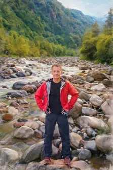 Мужской портрет под масло. На картине изображён мужчина в красной куртке, чёрной футболке и тёмных джинсах, стоящий на камнях у горной реки — он упёр руки в бока и смотрит прямо в камеру с лёгкой улыбкой. На заднем плане раскинулся живописный пейзаж: бурлящая река среди крупных валунов, густые зелёные леса и величественные горы, окутанные лёгкой дымкой. Художник Анна