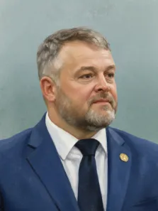 Мужской портрет в стиле под масло. На картине изображён мужчина средних лет с седеющими волосами и аккуратной бородой, одетый в строгий синий костюм, белую рубашку и тёмный галстук; на лацкане пиджака закреплён золотой значок. Фон выполнен в спокойных серо-голубых тонах, подчёркивающих официальность и сдержанность образа. Художник Ирина