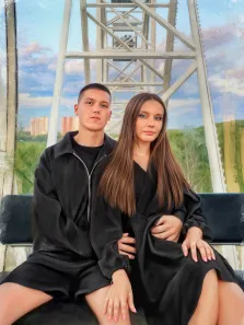 Парный портрет под масло. На картине изображены молодой мужчина и женщина, сидящие рядом на скамейке внутри конструкции колеса обозрения. Оба одеты в чёрное — мужчина в шортах и куртке-оверсайз, женщина в длинном платье; на фоне видны зелёные поля, городские здания и голубое небо с облаками. Художник Анна