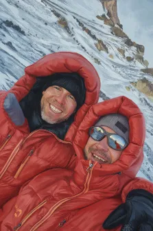 Парный портрет под масло. На картине изображены два альпиниста в ярко-красных пуховых куртках, улыбающихся и позирующих для селфи на заснеженном горном склоне. На заднем плане видны скалистые выступы и снежное покрывало, создающие ощущение высоты и суровой красоты горной природы. Художник Антонина