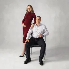 Парный портрет под масло. Беременная женщина в элегантном бордовом платье нежно кладёт руку на плечо сидящего рядом мужчины. Мужчина в светлой рубашке и чёрных брюках сидит на стуле, оба выглядят сдержанно и гармонично на нейтральном светлом фоне. Художник Антонина