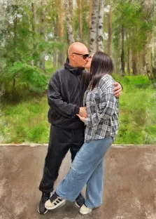 Парный портрет в стиле под масло. На картине изображена пара, целующаяся на фоне леса: мужчина в чёрной одежде и тёмных очках обнимает женщину в клетчатой рубашке и джинсах. Их поза передаёт теплоту и близость, а размытые зелёные оттенки леса создают спокойную, романтичную атмосферу. Художник Анна