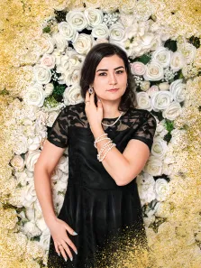 Женский портрет в стиле под масло. На картине изображена молодая женщина в элегантном чёрном платье с кружевными рукавами, украшенная жемчужными браслетами, серьгами и ожерельем. Она позирует на фоне пышной композиции из белых и кремовых роз, обрамлённой золотистыми блёстками, что придаёт образу торжественность и изысканность. Художник Артём