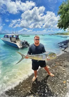 Мужской портрет в стиле под масло. На картине изображён мужчина в тёмной футболке и шортах, в солнечных очках, стоящий на галечном берегу и держащий в руках крупную серебристую рыбу. На фоне — бирюзовая морская гладь, лодка с удочками и яркое небо с пушистыми облаками, создающие атмосферу удачного рыболовного дня. Художник Анна