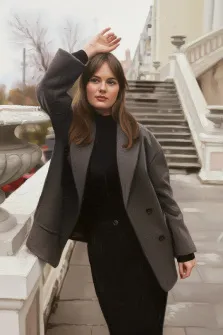 Женский портрет в стиле под масло. На картине изображена молодая женщина в элегантном чёрном платье и сером пальто, которая стоит на фоне парадной лестницы с балюстрадой и вазонами. Она слегка приподняла руку к голове, её взгляд спокоен и направлен вперёд, создавая ощущение уравновешенной задумчивости. Художник Анна