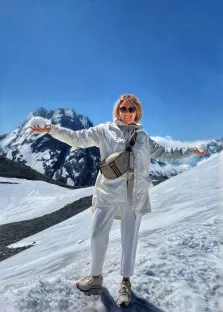 Женский портрет в стиле под масло.На картине изображена женщина в светлой одежде и с сумкой через плечо, стоящая на заснеженном склоне горы. Она улыбается, раскинув руки в стороны, в каждой руке держит снежок, на фоне видны величественные заснеженные вершины и ярко-голубое небо. Художник Александра