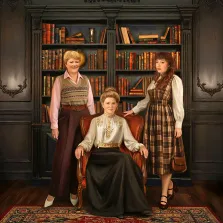 Семейный портрет в стиле под масло. На картине изображены три женщины в интерьере библиотеки: пожилая дама в элегантном наряде с золотой вышивкой сидит в кресле в центре, по бокам стоят две другие женщины — одна в свитере и длинной юбке, другая в клетчатом платье с сумкой. На заднем плане — массивный книжный шкаф, заполненный книгами, и узорчатый ковёр на полу, создающие атмосферу уюта и респектабельности. Художник  Анастасия