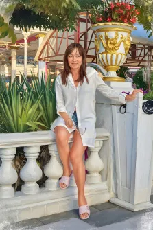 Женский портрет в стиле под масло. На картине изображена женщина в светлой рубашке и розовых тапочках, сидящая на белом балюстраде и небрежно опирающаяся рукой на декоративный вазон с яркими красными цветами. На фоне видны зелёные растения, архитектурные элементы и вывески, создающие атмосферу солнечного, уютного места на открытом воздухе. Художник Анна