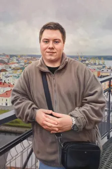 Мужской портрет в стиле под масло. На картине изображён молодой человек в коричневой куртке, с чёрной сумкой через плечо и наручными часами, стоящий на фоне городского пейзажа. За его спиной видны разноцветные здания, портовые краны и пасмурное небо, создающие атмосферу спокойного, немного задумчивого дня. Художник Антонина