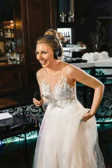 Женский портрет под масло. На портрете изображена улыбающаяся женщина в белом свадебном платье с кружевными деталями, держащая в руке микрофон. На голове у неё крупные наушники, она выглядит радостной и энергичной, словно выступает на сцене в уютном зале с барной стойкой на заднем плане. Художник: Ирина