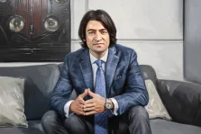 Мужской портрет под масло. На портрете изображён мужчина с длинными тёмными волосами, одетый в элегантный синий клетчатый костюм с галстуком, сидящий на тёмном диване с подушками. Его руки сложены перед собой, взгляд спокоен и сосредоточен, а на заднем плане видна картина с изображением передней части старинного автомобиля. Художник: Анастасия