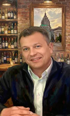 Мужской портрет под масло. На портрете изображён улыбающийся мужчина средних лет в тёмной куртке поверх светлой рубашки, сидящий в расслабленной позе с сложенными руками. Фон композиции — интерьер бара с кирпичной стеной, полками, уставленными бутылками вина, и картиной с изображением сталинского небоскрёба в золотой раме. Художник: Ирина