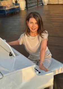 Женский портрет под масло. На портрете изображена женщина в светлом свободном наряде, сидящая за белым роялем — она слегка наклонена вперёд, с тёплой улыбкой смотрит прямо на зрителя. Мягкий солнечный свет подчёркивает её расслабленную позу и создаёт атмосферу уюта и гармонии, а фон с деревянной террасой и мебелью добавляет композиции лёгкую изысканность. Художник: Ирина