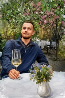 Мужской портрет под масло. На портрете изображён молодой мужчина в тёмно-синей куртке, сидящий за столом с бокалом белого вина в руке; его взгляд устремлён вверх, на лице — лёгкая улыбка. На столе перед ним стоит ваза с полевыми цветами, а на заднем плане виднеется уютная веранда, окружённая цветущими деревьями. Художник: Анна 