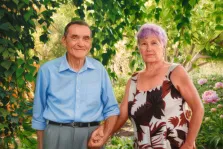 Парный портрет под масло. На портрете изображена пожилая пара: мужчина в светло-голубой рубашке и серых брюках с тёплой улыбкой держит за руку женщину с короткими сиреневыми волосами, одетую в платье с крупным цветочным принтом. Они стоят на фоне зелёной листвы и цветущих пионов, что создаёт атмосферу уюта и гармонии. Художник: Александра