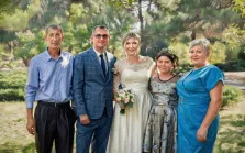 Семейный портрет под масло. На портрете изображена семья из пяти человек на фоне зелёного парка: в центре — невеста в белом кружевном платье с букетом, рядом с ней — жених в синем клетчатом костюме с галстуком; по бокам стоят двое мужчин и две женщины в нарядных платьях, все выглядят радостными и собранными для торжественного момента. Композиция передаёт атмосферу праздника и семейного единства, а мягкий дневной свет подчёркивает теплоту сцены. Художник: Ирина