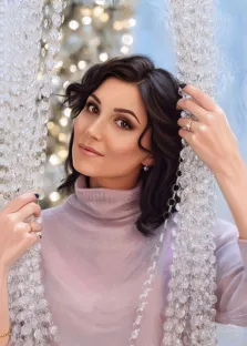 Женский портрет под масло. На портрете изображена женщина с тёмными волнистыми волосами, одетая в нежный светло-розовый свитер с высоким воротом; она слегка улыбается и держит в руках декоративные цепочки из прозрачных бусин, создавая ощущение лёгкости и элегантности. Фон размытый, с мягкими световыми бликами, напоминающими новогодние огоньки, что придаёт изображению праздничную и утончённую атмосферу. Художник: Александра