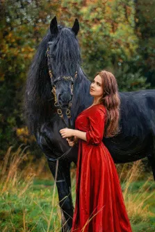 Женский портрет под масло. На портрете изображена молодая женщина в длинном ярко-красном платье, нежно держащая поводья величественной чёрной лошади с густой гривой и изысканной золотой сбруей. Фон композиции — размытый осенний пейзаж с зелёно-жёлтой листвой и высокой травой, создающий атмосферу гармонии между человеком и природой. Художник: Ирина