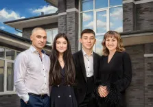 Семейный портрет под масло. На портрете изображена семья из четырёх человек — мужчина в светлой рубашке и тёмных брюках, молодая девушка в чёрном платье с длинными волосами, юноша в чёрном костюме и белой рубашке, а также женщина в чёрном наряде с перьевыми манжетами. Они стоят на фоне современного дома с большими окнами, все смотрят в камеру с лёгкими улыбками, создавая ощущение тепла и единства. Художник: Анна 