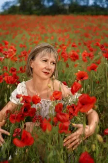 Женский портрет под масло. На портрете изображена женщина с короткими светлыми волосами в лёгкой белой блузе, стоящая среди густого макового поля; её взгляд устремлён в сторону, а на лице — спокойная, чуть задумчивая улыбка. Яркие красные маки окружают её со всех сторон, создавая контраст с её светлым нарядом и подчёркивая гармонию между человеком и природой. Художник: Ирина