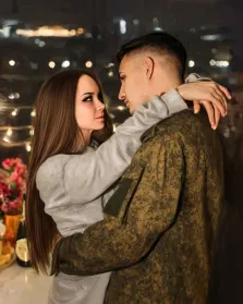 Парный портрет под масло. На портрете изображены молодой мужчина в камуфляжной форме и девушка с длинными каштановыми волосами в светло-сером свитере; они стоят в тесных объятиях, глядя друг другу в глаза с нежностью и трепетом. Размытый фон с огнями ночного города и элементами праздничного стола создаёт атмосферу тёплого, интимного момента встречи. Художник: Анастасия 