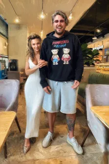 Парный портрет под масло. На портрете изображены мужчина и женщина, позирующие вместе в интерьере кафе: мужчина в тёмной худи с рисунком медвежат, светлых шортах и белых кроссовках, с татуировкой на ноге; женщина — в длинном белом платье и блестящих туфлях, с объёмной причёской, нежно улыбается, держась за руку мужчины. Художник: Александра 