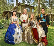 Семейный портрет в образе. На картине изображён семейный портрет в исторических образах: две женщины в роскошных придворных платьях с коронами и двое мужчин в парадных военных мундирах с орденами. Композиция размещена на фоне деревянного дома, окружённого зелёными деревьями, что создаёт контраст между царственной элегантностью персонажей и спокойной природной обстановкой. Художник Анастасия