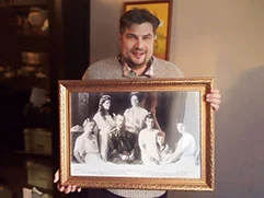 Мужчина держит в руках подарок от своего друга — картину с изображением семьи императора Николая II, обрамленную в багетную раму.