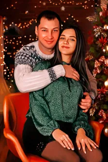 Парный новогодний портрет стилизован Под масло, молодой человек в белом вязаном свитере обнимает за плечи девушку в зелёной блузке, пара сидит возле новогодней ёлки, художник Юлия 