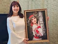 Женщина в белом платье держит в руках подарок — портрет, выполненный по фотографии на холсте, где она изображена в народной одежде у березы