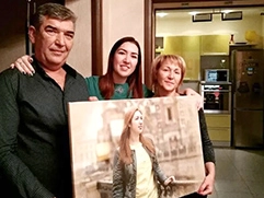 Три человека: мужчина, женщина и молодая девушка; они стоят в квартире, на заднем плане виден кухонный интерьер с холодильником и шкафчиками; они держат в руках большой портрет; на портрете изображена та же девушка, что и на переднем плане, на картине она стоит на улице на фоне собора Спас на Крови в СПб, одетая в светлую кофту и куртку; портрет выполнен в акварельном стиле и напечатан на холсте