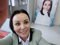 На фото изображена женщина, которая улыбается и позирует, делая селфи; на заднем плане виден её портрет на холсте, выполненный в стиле дрим-арт