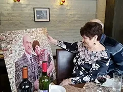 На фото изображена женщина, сидящая за праздничным столом; она держит в руках большую фотомозаику с изображением мужчины и женщины; картина напечатана на холсте по фотографиям на заказ ей в подарок