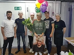 В центре фото женщина держит подарок от коллектива на день рождения коллеги — её портрет на холсте в стиле гранж; вокруг неё стоят ещё пять человек, одетых в рабочую одежду, включая поварские халаты и сетки для волос, видны разноцветные воздушные шары и цветы, что создаёт праздничное настроение