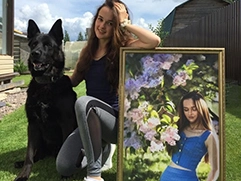 На фото изображена девушка, сидящая на траве рядом с черной собакой; обстановка вокруг — двор с зеленью и ясным небом; девушка держит в руках подарок — портрет на холсте, обрамленный в багет, на котором изображена она же, окруженная цветами, в стиле под масло.